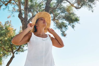 Sahil kenarında hasır şapka takan çekici orta yaşlı bir kadın. Mutlu olgun kadın denizde özgürlüğün tadını çıkarıyor ve gökyüzüne bakıyor..