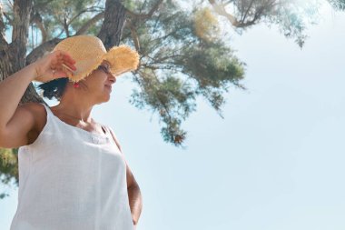 Sahil kenarında hasır şapka takan çekici orta yaşlı bir kadın. Mutlu olgun kadın denizde özgürlüğün tadını çıkarıyor ve gökyüzüne bakıyor..