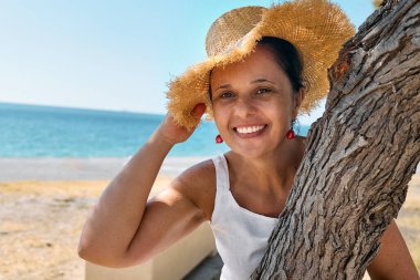 Sahil kenarında hasır şapka takan çekici orta yaşlı bir kadın. Mutlu olgun kadın denizde özgürlüğün tadını çıkarıyor, ağaca sarılıyor ve kameraya bakıyor..