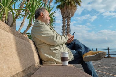 Gülümseyen orta yaşlı sakallı adam güneşli bir kış ya da bahar günü sahilde bankta otururken akıllı telefon kullanıyor..