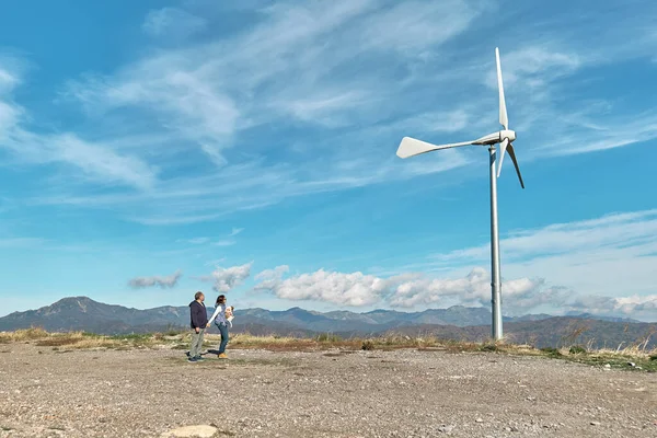 Green energy concept. Couple with dog staring the wind turbine for generating electricity in wind farm or wind park while walking near mountain top industrial wind power plant.