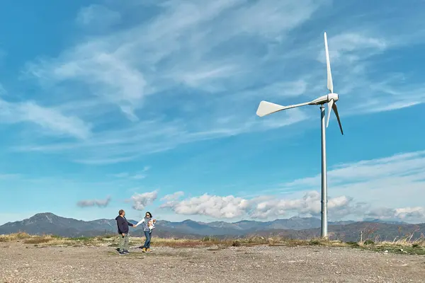 Green energy concept. Couple walking with dog near mountain top industrial wind power plant, wind turbine for generating electricity in wind farm or wind park.
