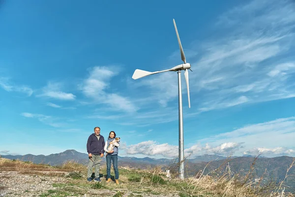 Green energy concept. Couple with dog near mountain top industrial wind power plant, wind turbine for generating electricity in wind farm or wind park.