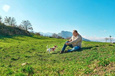 Gri saçlı adam dağlık bölgede çayırda Jack Russell köpeğiyle yürüyüşe çıkıyor. Olgun bir adam köpeğiyle dışarıda oynuyor..