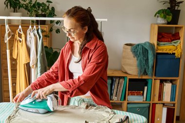 Evde ütü masasında çamaşır ütüleyen mutlu orta yaşlı kadın. Günlük ev hanımı rutini. Oda servisi kavramı.