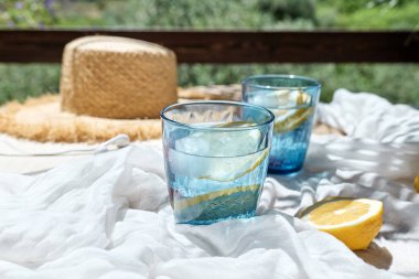 Yaz ferahlatıcı limonata ya da limon dilimli alkolsüz kokteyl. Bahçedeki beyaz battaniyede içilen bardaklar. Taze, sağlıklı, soğuk limon suyu. Limonlu su. Yaz havası.