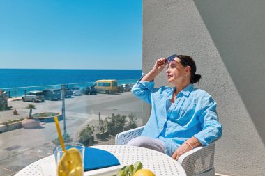 Tatmin olmuş olgun bir kadın yazın otel balkonunda dinleniyor ve denizi düşünüyor. Akdeniz 'de dinlenme ve yaz tatili kavramı.
