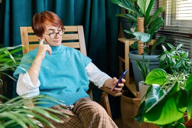 Balkonda akıllı telefon kullanan bir kadın ve sonbahar günü evde yeşil ev bitkileri var. Modern yaşam tarzı, bağlantı, blog yazma, iş dünyası, serbest çalışma.