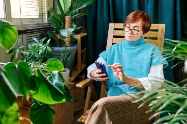 Balkonda akıllı telefon kullanan bir kadın ve sonbahar günü evde yeşil ev bitkileri var. Modern yaşam tarzı, bağlantı, blog yazma, iş dünyası, serbest çalışma.