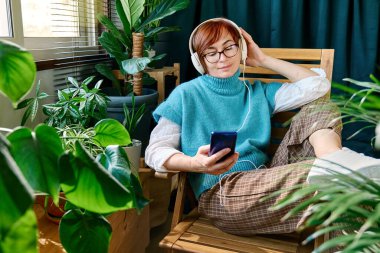 Müzik dinleyen bir kadın ya da sonbahar günü yeşil ev bitkileriyle birlikte balkonda kulaklıkla akıllı telefon uygulamasından podcast yapan bir kadın. Modern yaşam tarzı, bağlantı, blog yazma, iş dünyası, serbest çalışma.