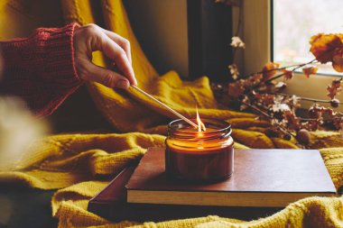 Yanan kibritle birlikte pencere pervazında mum yakıyor. Balkabaklarıyla rahat bir sonbahar, yün yün ekose örgü ve kitaplarla dolu. Sonbahar evi dekorasyonu. Samimi düşüş havası. Şükran Günü. Cadılar Bayramı