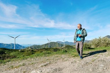 Dürbün kullanan erkek turist rüzgar türbininin önünde dağ manzaralı bir rüzgar çiftliğinde.