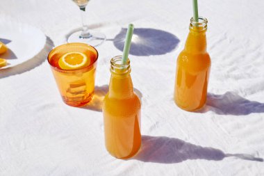 Soğuk yaz içkisi. Masada beyaz masa örtüsüyle pipet içerek taze portakal suyunu şişelerde tazeliyorum..