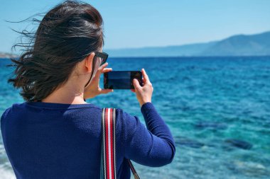Rüzgarlı bir bahar günü sahilde akıllı telefonuyla fotoğraf çeken turist kadının arka görüntüsü..