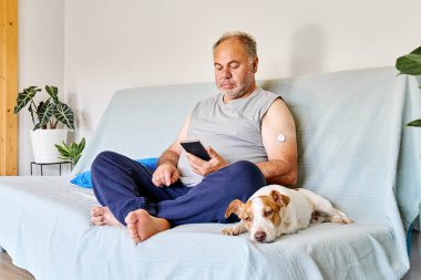 Kolunda şeker hastası ve devamlı glikoz monitörü olan olgun bir adam sağlığını kontrol etmek için mobil uygulama kullanırken aynı zamanda evinde Jack Russell Terrier 'ın köpeği ile kanepede oturuyor.