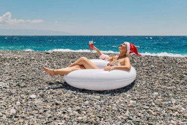 Güzel, bronz tenli, kırmızı Noel Baba şapkası takan ve zebra bikinili bir kadın plajda kırmızı içecekle büyük beyaz bir yüzme ringinde dinleniyor. Sıcak ülkelerde Noel tatili.