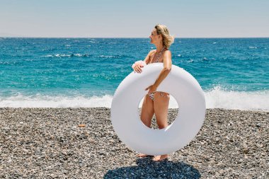 Bikini giyen sarışın kadın plajda beyaz, şişme yüzük taşıyor.