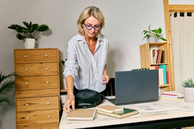 Düşünceli sarı saçlı iş kadını iş yerindeki masasından not defteri alıyor.