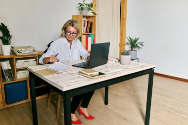 Sarı saçlı iş kadını, gözlüklü, ofiste dizüstü bilgisayara odaklanmış.