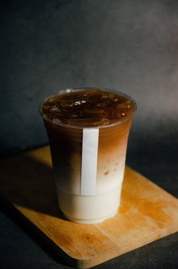 Plastik camla kaplanmış latte, beyaz etiketli, tahta plakalı, gri arka planlı.