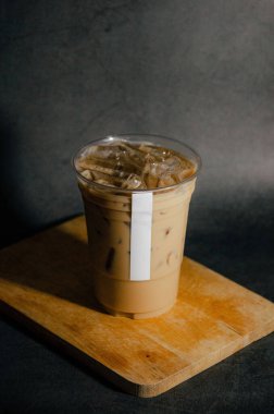 Buzlu kahveyi plastik bardakta, beyaz etiketli, tahta tabakta, gri arka planda.