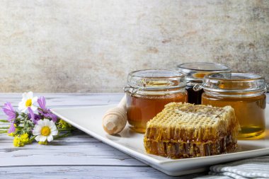 Bal peteğinin yanında farklı bal türleri olan kavanozlar ve yabani çiçeklerle dolu bir tabakta bal kapanı.