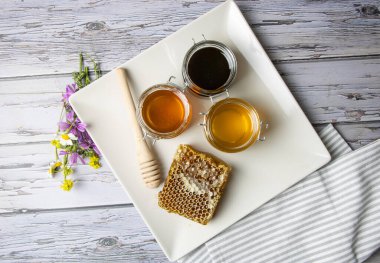Bal peteğinin yanında farklı bal türleri olan kavanozlar ve yukarıdan görülen yabani çiçeklerle dolu bir tabakta bal kapsülü.