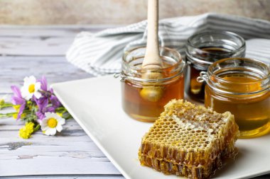 Bal peteğinin yanında farklı bal türleri olan kavanozlar ve yabani çiçeklerle dolu bir tabakta bal kapanı.