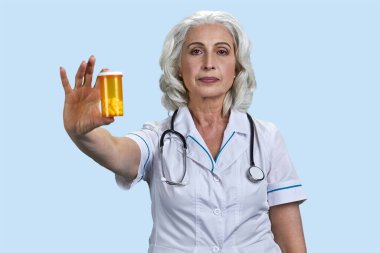 Sarı ilaç şişesini gösteren yaşlı bir kadın doktor. Pastel mavi arkaplanda izole edilmiş.