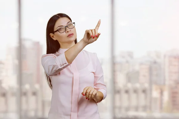 Young asian woman is pointing at invisible virtual. Businesswoman interact with invisible virtual screen by her index finger. Blurred windows background.