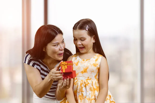 Young mother is open red gift box oh her surprised little daughter. Birthday present for little daughter.