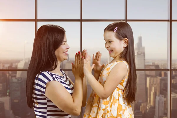 Side view of woman and her daughter are playing clapping game. Checkered window background with cityscape view.