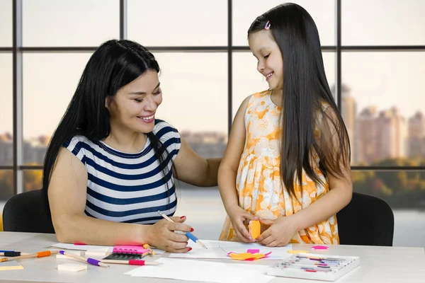 Mother and her daughter are drawing and making paper cutouts. Checkered window background with cityscape view.
