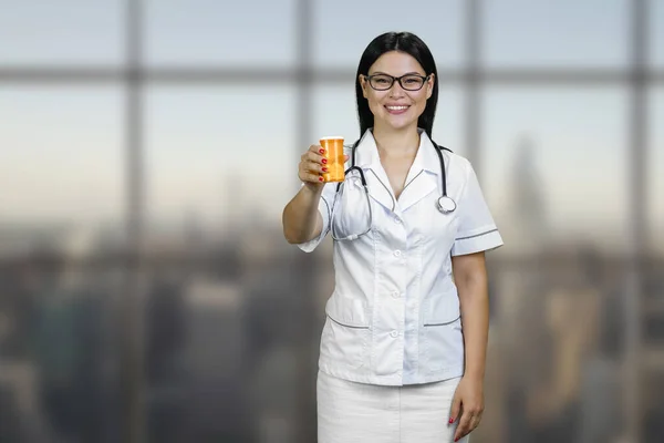 Portrait of a young female asian doctor is showing medicine can. Smiling cheerful brunette physician with glasses and stethoscope.