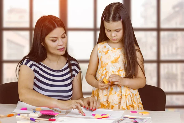Mother and her daughter are making paper cutouts. Blurred checkered window background.
