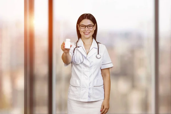 Young female asian doctor is advertising blank medicine bottle. Wearing glasses and stethoscope. Blurred windows background with evening sulight.