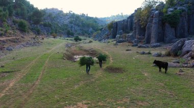 Landscape with majestic geological formations and people carrying branches through green field. Picturesque countryside view.
