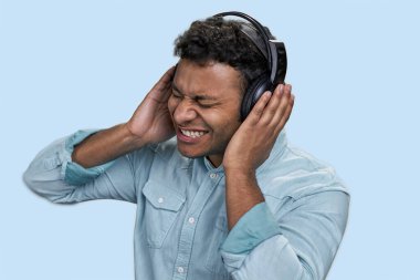 İfade veren mutlu Hintli genç adam mavi gömlek giyiyor kulaklıkla müzik dinliyor. Soluk mavi arkaplanda izole.