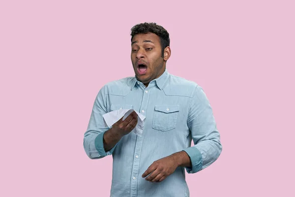 Sick ill young indian man sneezing or coughing by covering his face and nose with napkin. Isolated on pink background.