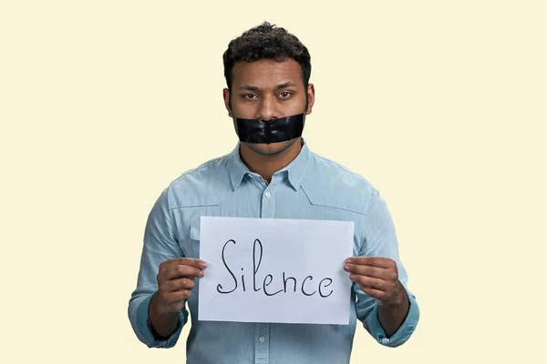 Indian man with mouth taped holding paper sheet with silence inscription. Isolated on beige background.