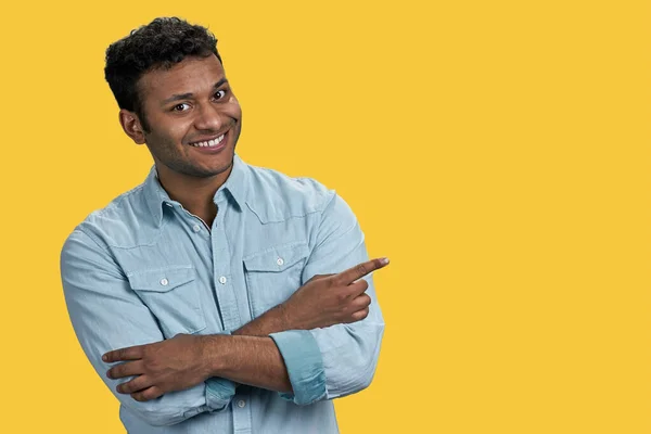 Indian man with folded arms pointing at copy space on the right. Isolated on vivid yellow background.