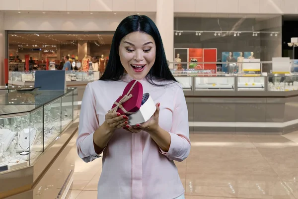 年轻的黑发亚洲女人打开一个心形礼品盒 惊讶的面部表情 背景中的商场内部 — 图库照片