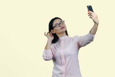 Gözlüklü sevimli Asyalı kadın akıllı telefonuyla selfie çekiyor. Sarıda izole edilmiş..