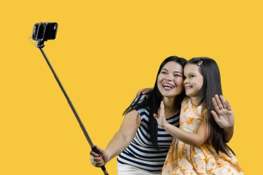 Anne ve kızı akıllı telefondan selfie çubuğu kullanarak ortak bir fotoğraf çekiyorlar. Sarı arkaplanda izole edilmiş.