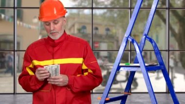 Kırmızı üniformalı bir inşaat işçisinin portresi para sayıyor ve parmağını kaldırıyor. Arka planda sokak manzaralı kapalı bir pencere..