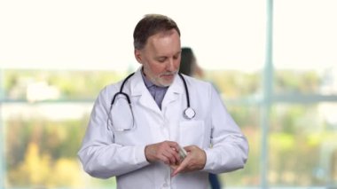 Mutlu kıdemli doktor parasını ceketinin cebine koyar ve kollarını kavuşturur. Arkaplanda bulanık pencereler.