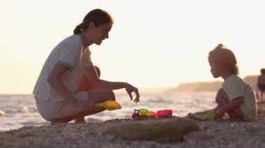 Küçük kız ve annesi kumsalda oyuncak ve kumla oynuyorlar. Arka planda akşam deniz manzarası.
