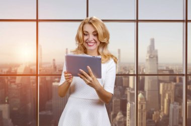 Gülümseyen orta yaşlı, beyaz, elinde dijital tablet olan sarışın kadın. Çevrimiçi sosyal medya haberleri okuyor. Akşam gün batımlı şehir manzarası.