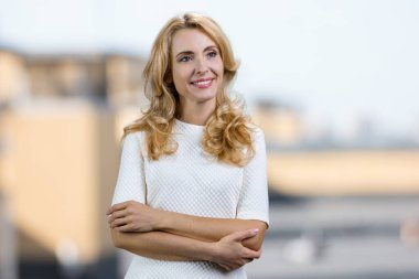 Çekici, düşünceli, orta yaşlı, kolları bağlı bir kadın. Arka planda bulanık şehir manzarası.