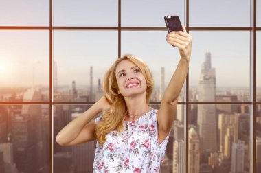 Gülümseyen iş kadını akıllı telefondan selfie çekiyor. Arkaplanda şehir manzarası olan işaretlenmiş pencere.